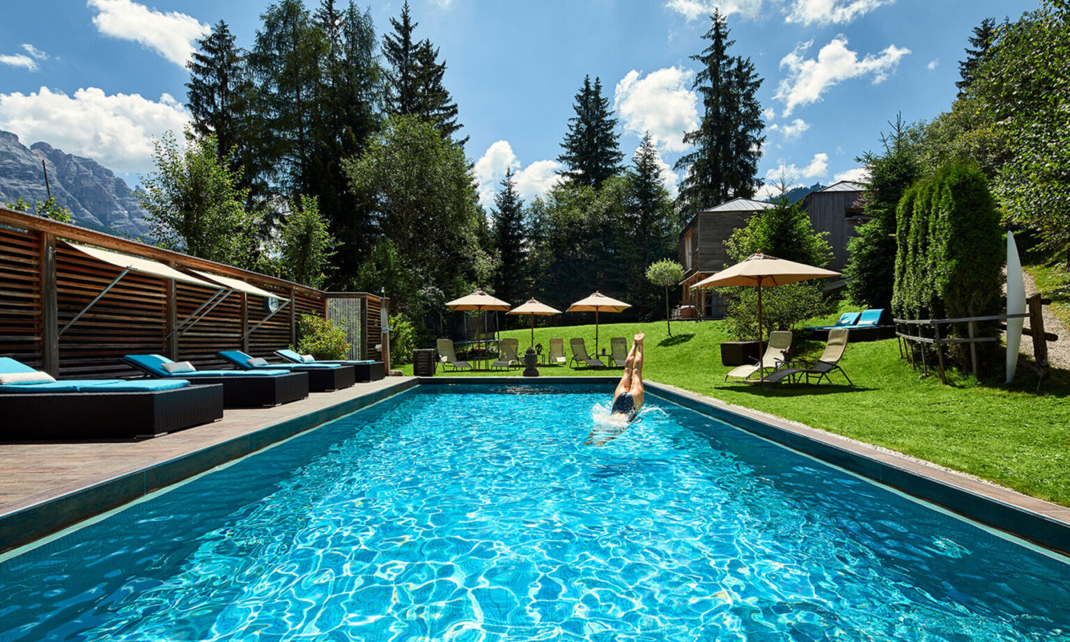 A person diving into a crystal-clear swimming pool surrounded by lush greenery, sun loungers, and umbrellas, with mountains in the background under a bright blue sky. Ideal for relaxation and outdoor enjoyment. | Only on SopranoVillas