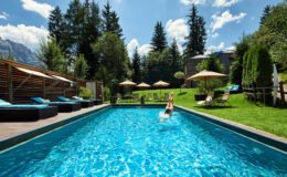 A person diving into a crystal-clear swimming pool surrounded by lush greenery, sun loungers, and umbrellas, with mountains in the background under a bright blue sky. Ideal for relaxation and outdoor enjoyment.