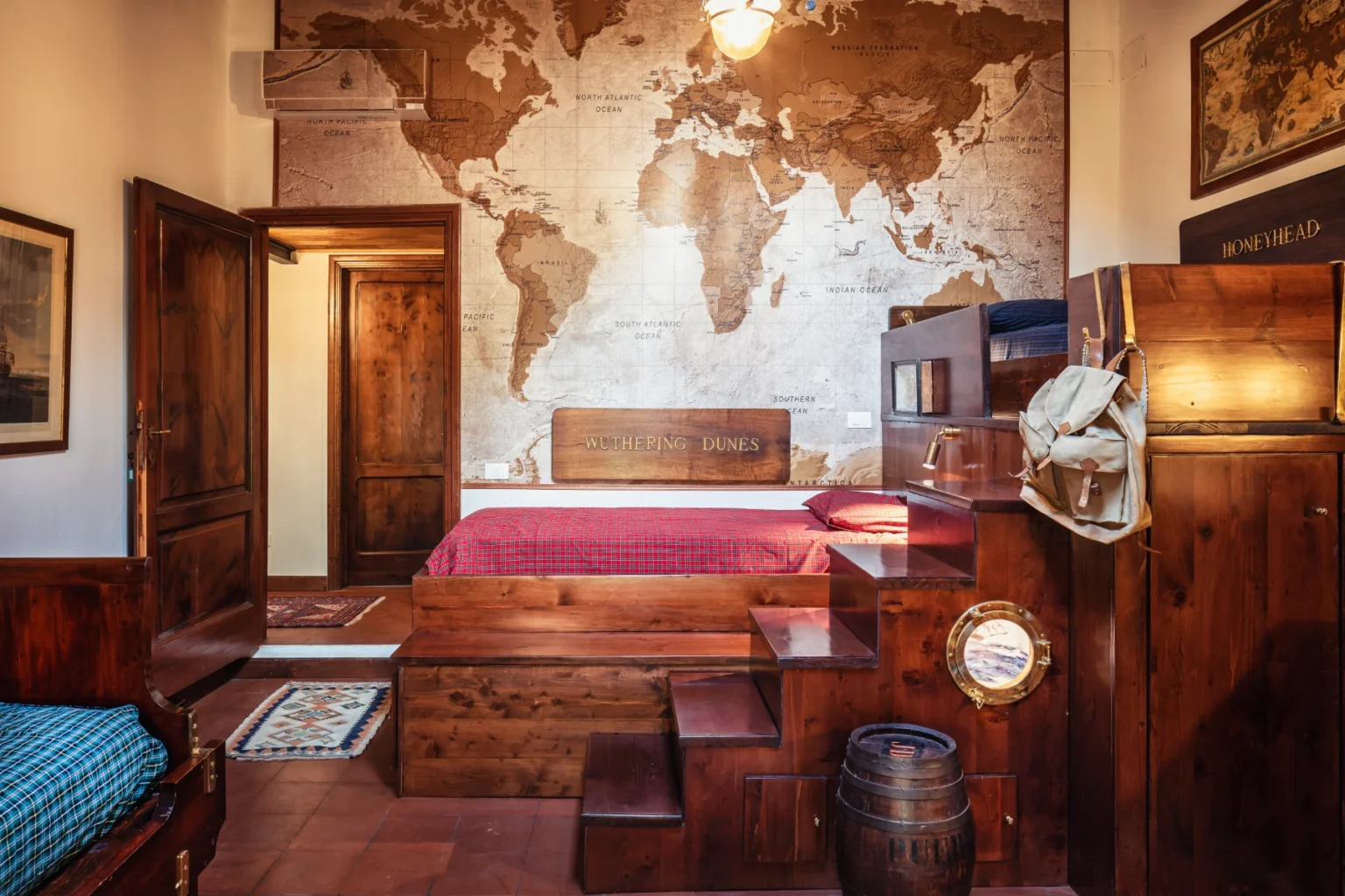 Cozy bedroom featuring a large world map mural, wooden furniture, a red bedspread, and decorative elements like a barrel and a vintage-style lamp. The room is warmly lit and has a rustic charm, ideal for travelers and explorers. | Only on SopranoVillas