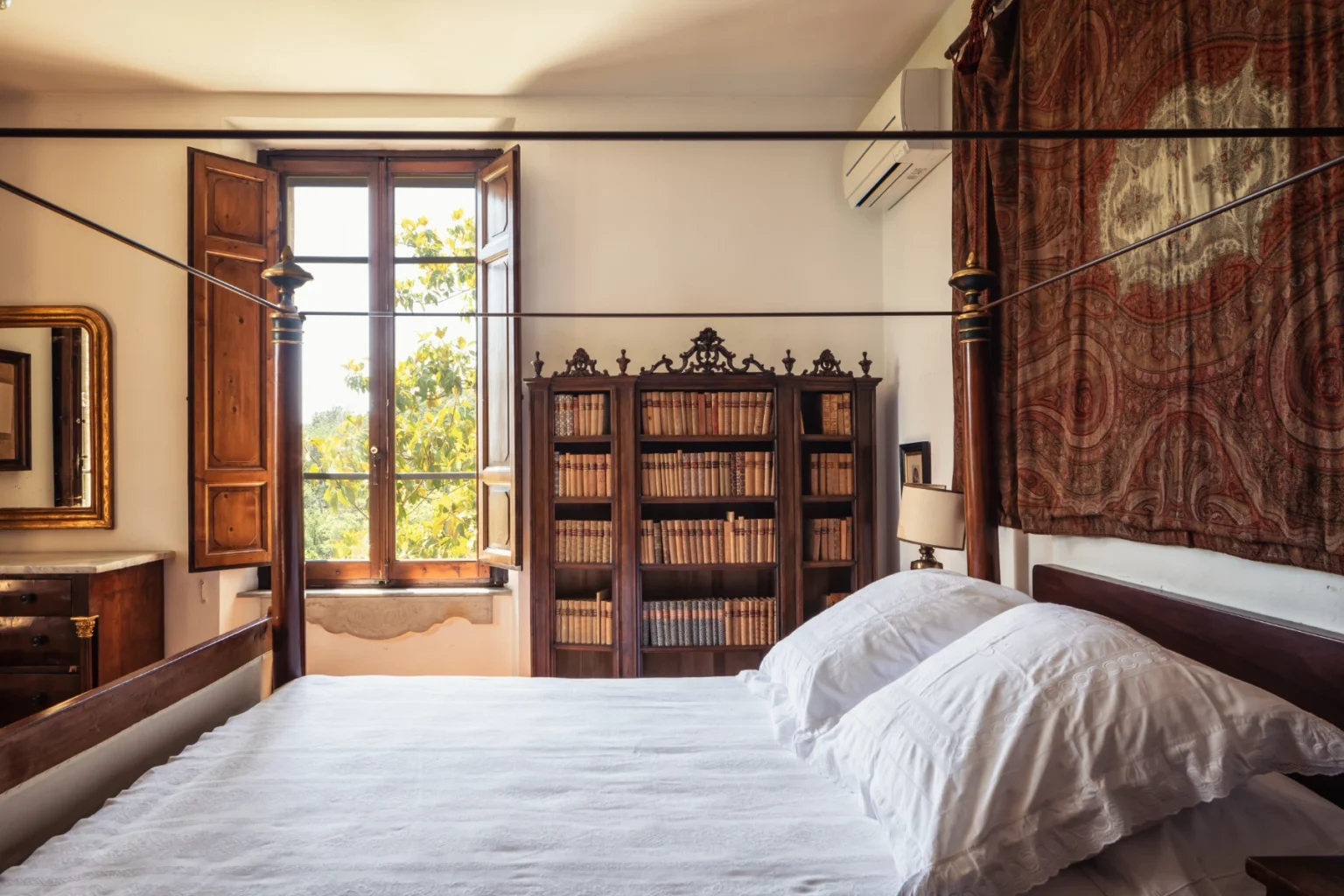 Cozy bedroom featuring a four-poster bed with white bedding, a wooden bookshelf filled with books, and large windows allowing natural light and a view of greenery. | Only on SopranoVillas
