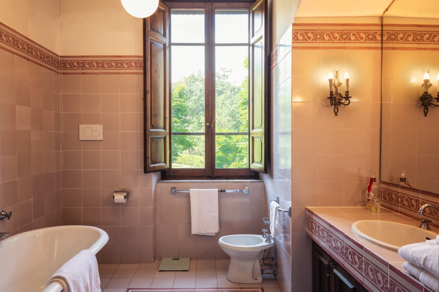 Spacious bathroom featuring a vintage bathtub, modern toilet, and elegant sink with a large mirror, complemented by natural light from an open window overlooking greenery. Decorative wall tiles and stylish sconces enhance the aesthetic appeal. | Only on SopranoVillas