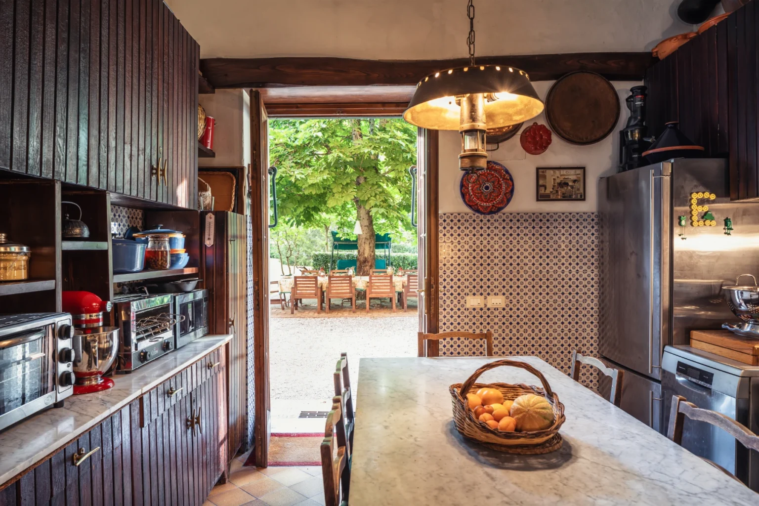 Modern kitchen interior with wooden cabinets, marble countertop, and a basket of fruits, featuring a door that opens to an outdoor dining area surrounded by greenery. | Only on SopranoVillas