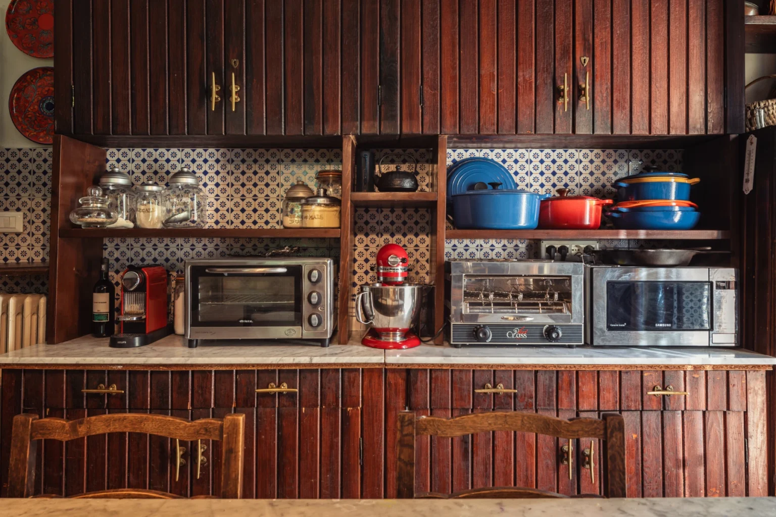 A cozy kitchen featuring wooden cabinetry, a countertop with various kitchen appliances including a toaster, mixer, and microwave, complemented by colorful cookware and decorative tiles. | Only on SopranoVillas