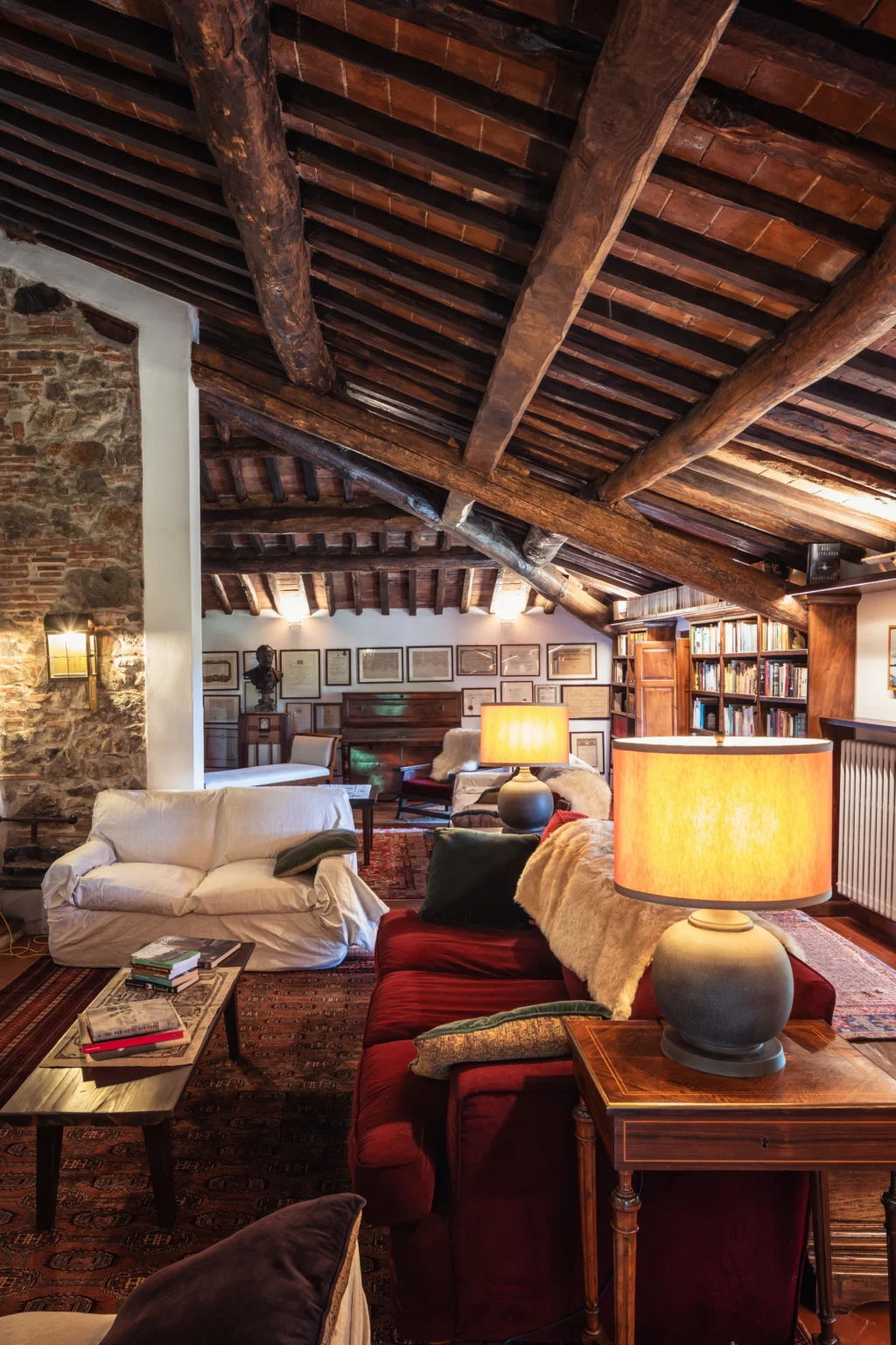 Cozy interior of a rustic living room featuring exposed wooden beams, elegant lamps, comfortable seating, and a wall adorned with framed artwork and bookshelves, creating a warm and inviting atmosphere. | Only on SopranoVillas