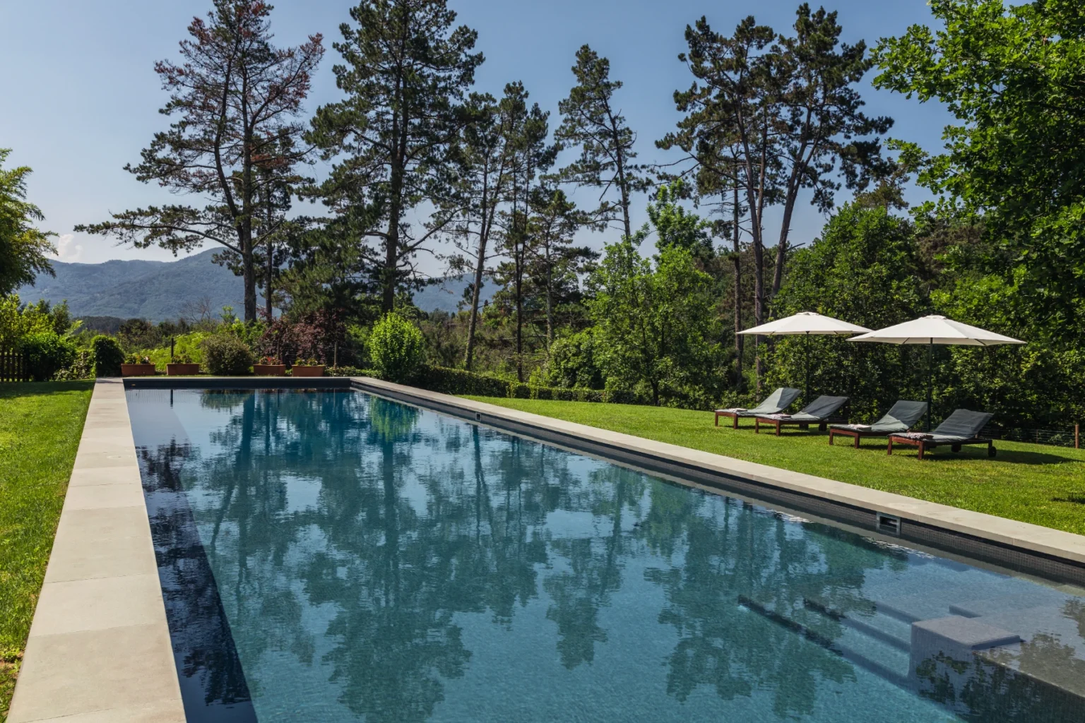 Luxury outdoor swimming pool surrounded by lush greenery and tall trees, featuring two sun loungers and shaded by white umbrellas, ideal for relaxation and leisure. | Only on SopranoVillas