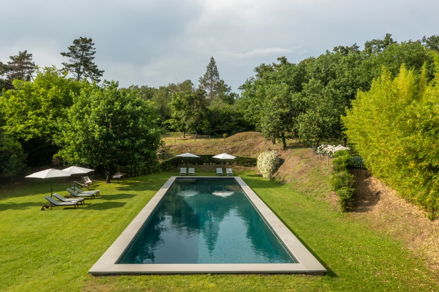 Serene outdoor pool surrounded by lush greenery and trees, featuring sun loungers and umbrellas, perfect for relaxation and leisure in a tranquil setting. | Only on SopranoVillas