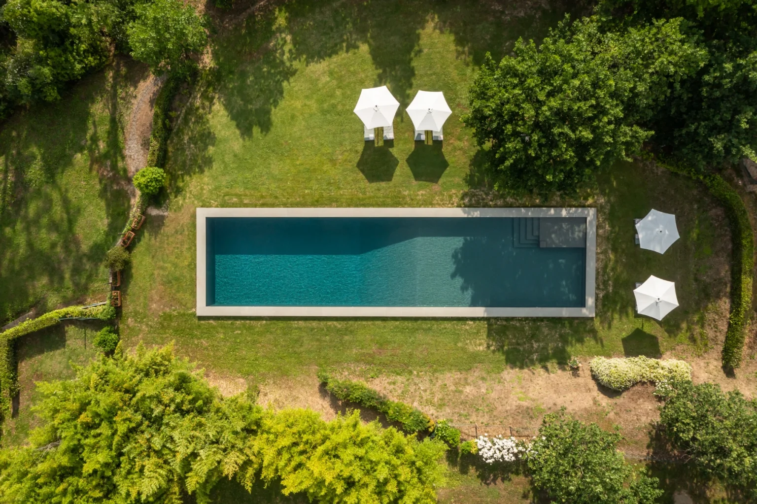 Aerial view of a modern rectangular swimming pool surrounded by lush green grass and trees, featuring two white umbrellas for shade. | Only on SopranoVillas