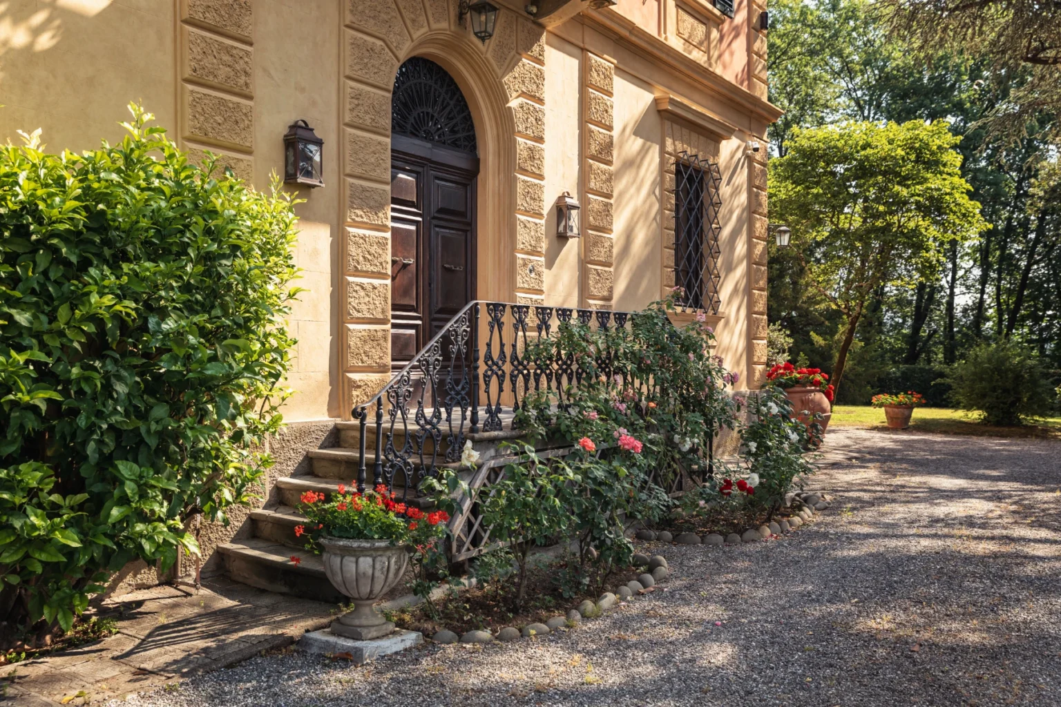 Elegant entrance of a villa featuring a wooden door, decorative wrought iron railing, and vibrant flower beds, surrounded by lush greenery and a gravel pathway. | Only on SopranoVillas