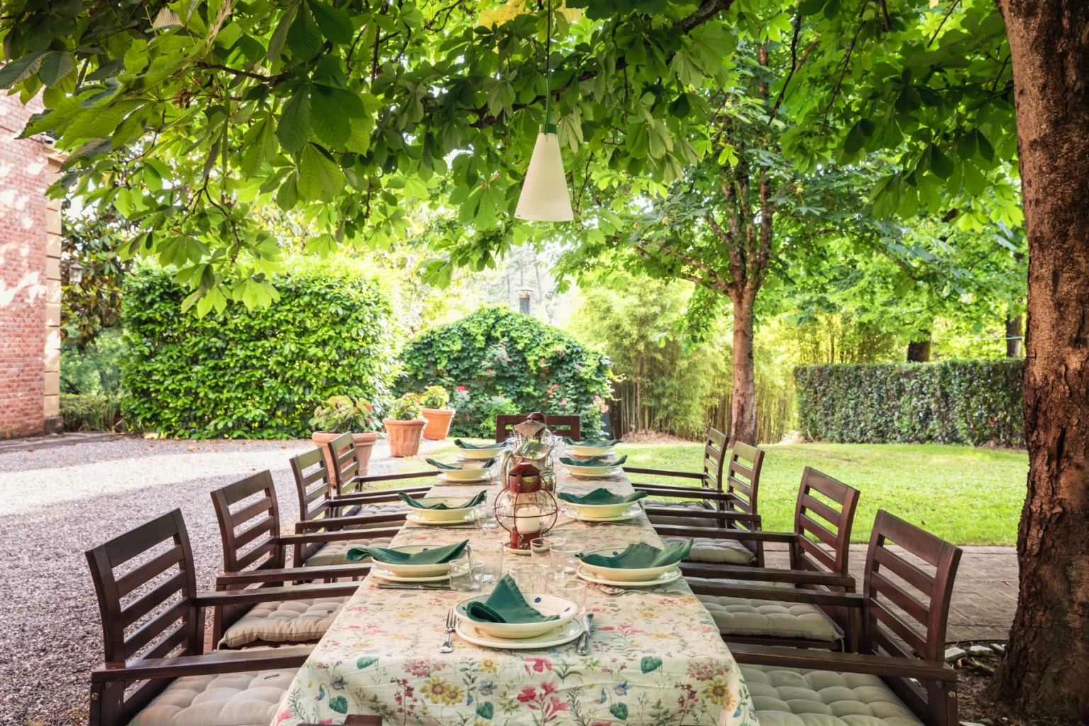 Outdoor dining setup under lush greenery featuring a long table with elegant tableware, surrounded by trees and garden elements, ideal for alfresco gatherings and events. | Only on SopranoVillas