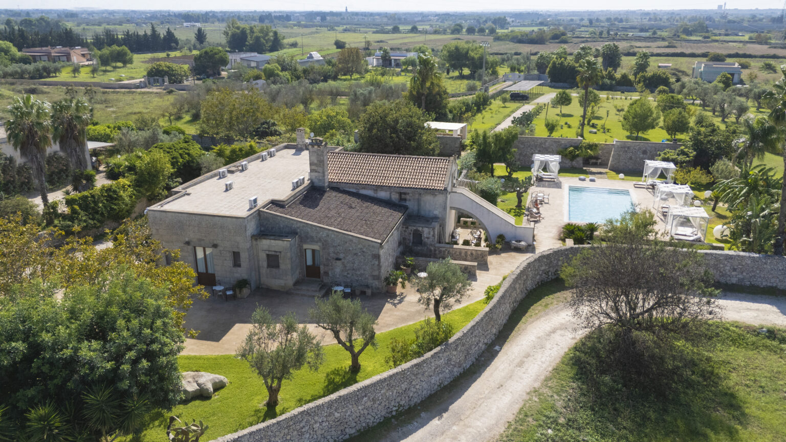 Aerial view of a luxurious stone villa surrounded by lush greenery, featuring a swimming pool and shaded lounging areas, set in a tranquil landscape with rolling hills in the background. | Only on SopranoVillas