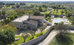 Aerial view of a luxurious stone villa surrounded by lush greenery, featuring a swimming pool and shaded lounging areas, set in a tranquil landscape with rolling hills in the background.