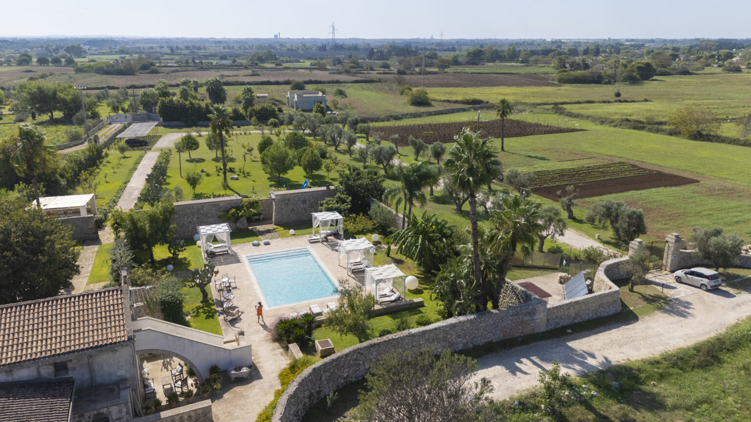 Aerial view of a serene countryside villa featuring a swimming pool surrounded by lush gardens, palm trees, and farmland, ideal for relaxation and outdoor activities. | Only on SopranoVillas