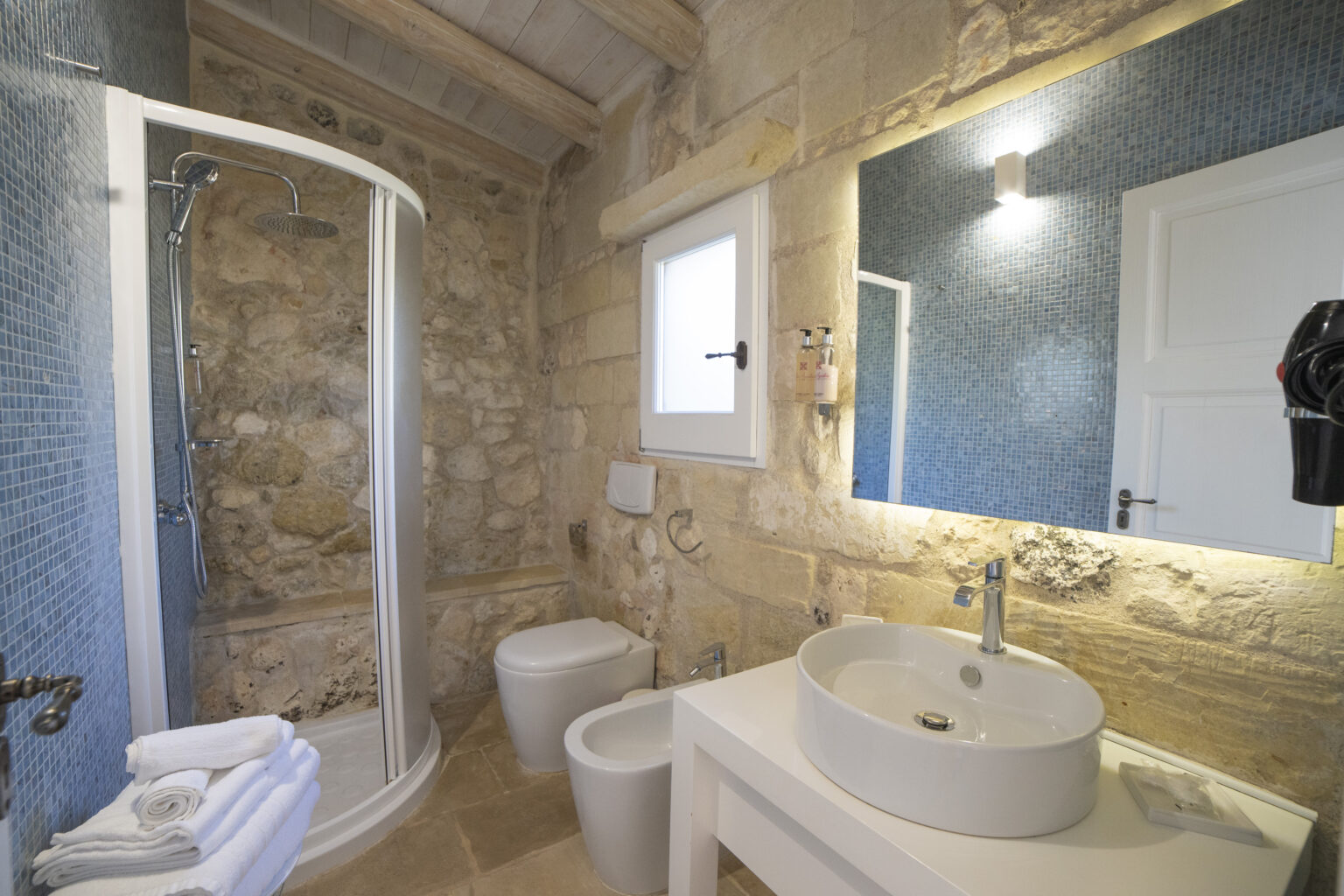Modern bathroom featuring a glass shower, stone walls, and a stylish sink with a mirror. The space is well-lit with natural light and includes white towels and a toilet. | Only on SopranoVillas