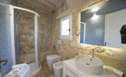 Modern bathroom featuring a glass shower, stone walls, and a stylish sink with a mirror. The space is well-lit with natural light and includes white towels and a toilet.