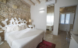 Cozy bedroom featuring a stone accent wall, a white bed with decorative pillow, a modern lamp, and a mirror. The room is illuminated by natural light through sheer curtains, showcasing a stylish interior with a plush area rug and contemporary furnishings. Ideal for a relaxing getaway.