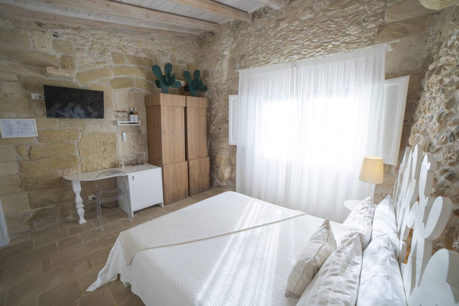 Cozy bedroom featuring a stone wall, a comfortable bed with decorative pillows, a minimalist desk, and a flat-screen TV. The room is illuminated by natural light filtering through sheer curtains, enhancing the inviting atmosphere. A wooden wardrobe and modern decor elements add to the charm of this relaxing space. | Only on SopranoVillas