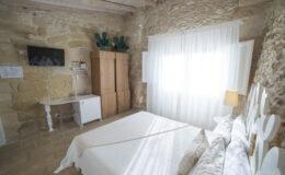 Cozy bedroom featuring a stone wall, a comfortable bed with decorative pillows, a minimalist desk, and a flat-screen TV. The room is illuminated by natural light filtering through sheer curtains, enhancing the inviting atmosphere. A wooden wardrobe and modern decor elements add to the charm of this relaxing space.