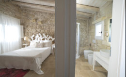 Cozy bedroom with stone walls, a large bed, and natural light, adjacent to a modern bathroom featuring a shower, toilet, and sink. Ideal for a relaxing getaway.