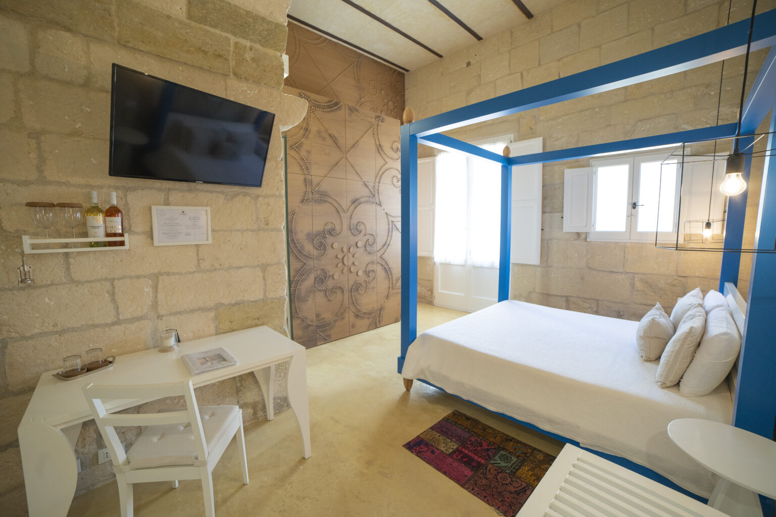 Cozy bedroom featuring a blue four-poster bed, stone walls, a flat-screen TV, and a stylish desk area. The room is illuminated by natural light from large windows, creating a warm and inviting atmosphere. | Only on SopranoVillas