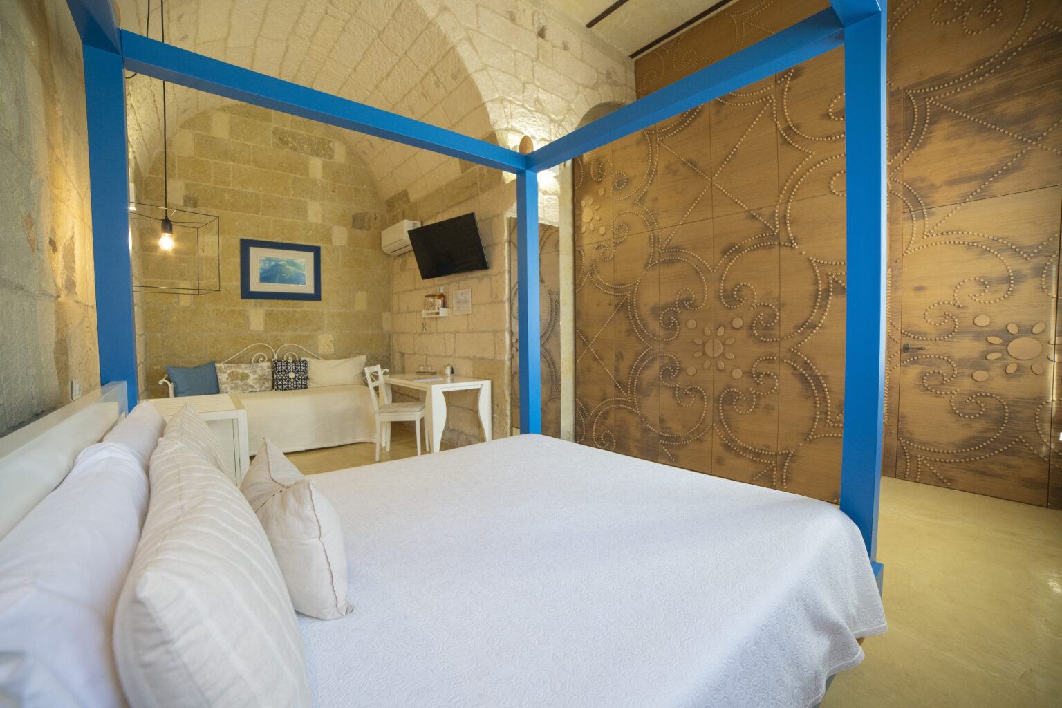 Cozy hotel room featuring a four-poster bed with white bedding, stone walls, and a blue accent frame. The space includes a comfortable seating area, a small dining table, and a wall-mounted TV, creating a welcoming atmosphere for relaxation. | Only on SopranoVillas