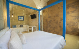 Cozy hotel room featuring a four-poster bed with white bedding, stone walls, and a blue accent frame. The space includes a comfortable seating area, a small dining table, and a wall-mounted TV, creating a welcoming atmosphere for relaxation.