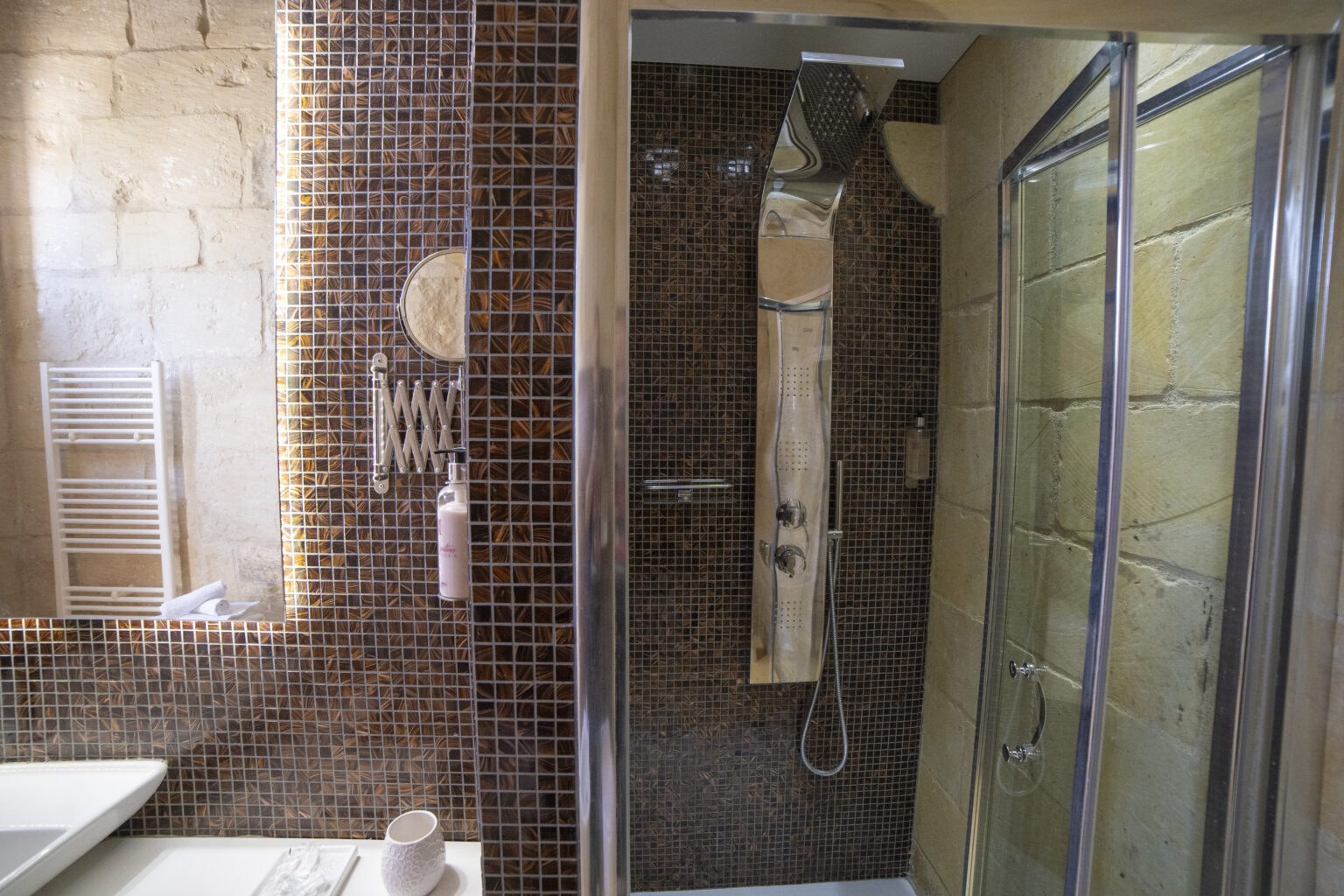 Modern bathroom featuring a stylish shower area with brown mosaic tiles, a sleek chrome shower panel, and a round mirror. Includes a white sink, a heated towel rack, and decorative accessories. | Only on SopranoVillas