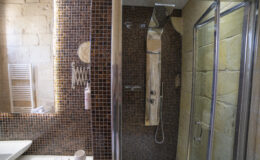 Modern bathroom featuring a stylish shower area with brown mosaic tiles, a sleek chrome shower panel, and a round mirror. Includes a white sink, a heated towel rack, and decorative accessories.