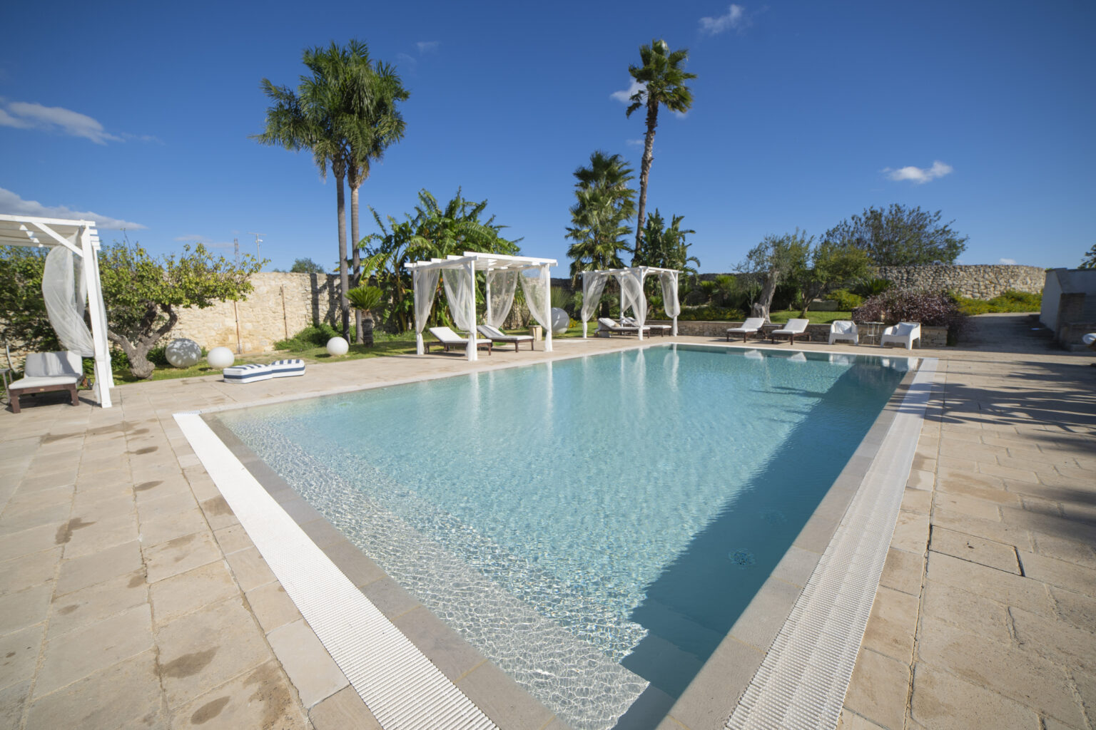 Luxury outdoor swimming pool surrounded by lush greenery and palm trees, featuring elegant cabanas and sun loungers, ideal for relaxation and leisure in a sunny setting. | Only on SopranoVillas