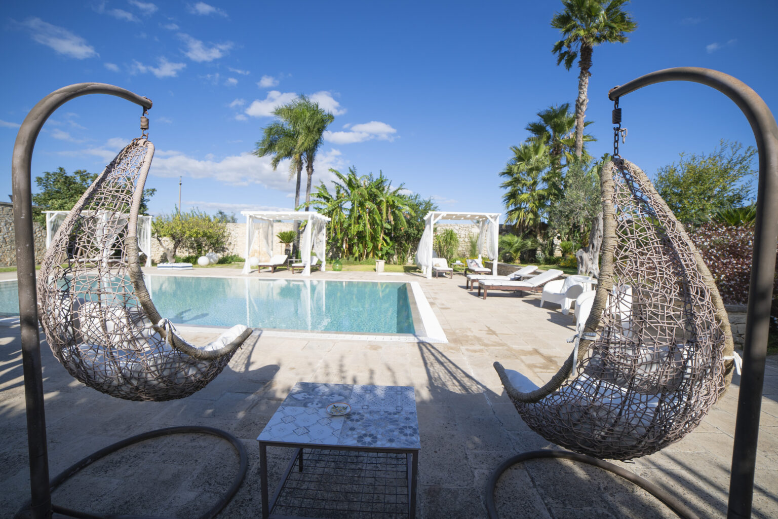 Outdoor oasis featuring a serene swimming pool surrounded by lush greenery and palm trees. Two stylish hanging chairs provide a cozy seating area, complemented by a small table. White sun loungers and cabanas create a relaxing atmosphere under a clear blue sky. Perfect for leisure and relaxation. | Only on SopranoVillas