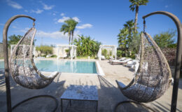 Outdoor oasis featuring a serene swimming pool surrounded by lush greenery and palm trees. Two stylish hanging chairs provide a cozy seating area, complemented by a small table. White sun loungers and cabanas create a relaxing atmosphere under a clear blue sky. Perfect for leisure and relaxation.