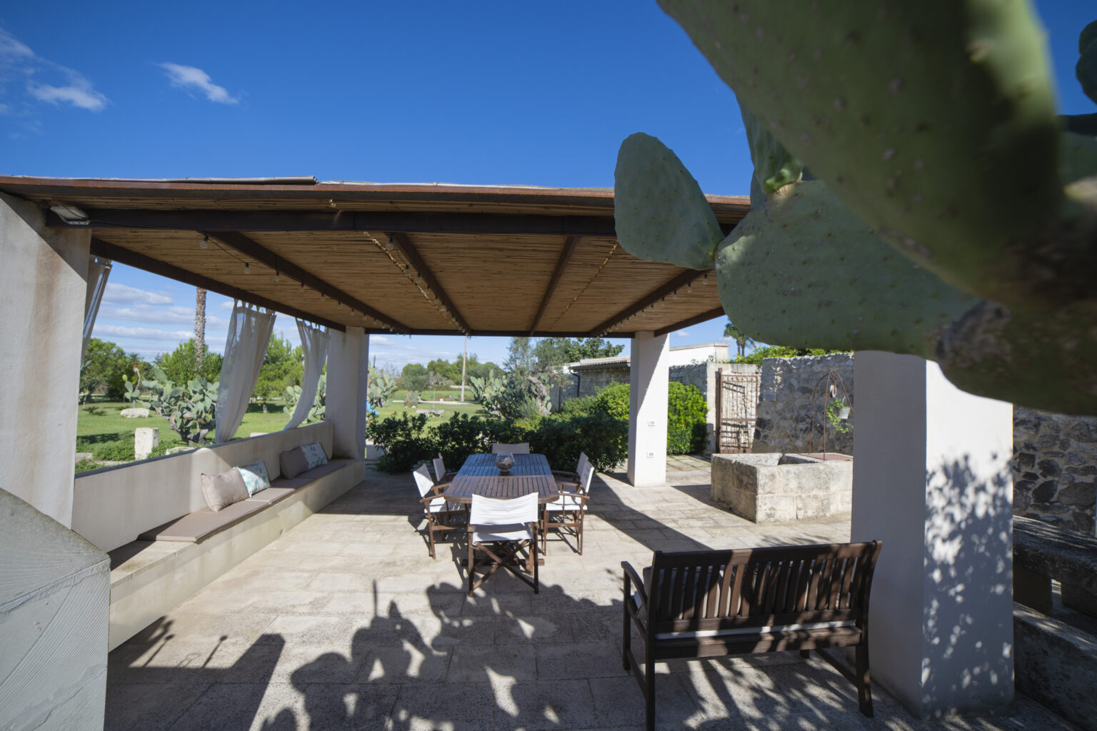 Outdoor patio with wooden roof, seating area, and stone walls surrounded by greenery and cacti under a clear blue sky. Ideal for relaxation and dining. | Only on SopranoVillas