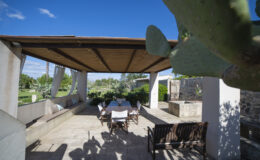 Outdoor patio with wooden roof, seating area, and stone walls surrounded by greenery and cacti under a clear blue sky. Ideal for relaxation and dining.