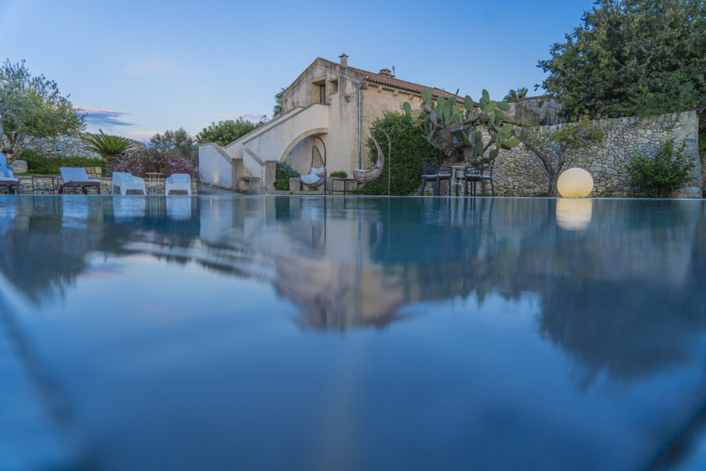 Serene outdoor pool reflecting a charming villa surrounded by lush greenery and modern seating, capturing a tranquil sunset ambiance. | Only on SopranoVillas