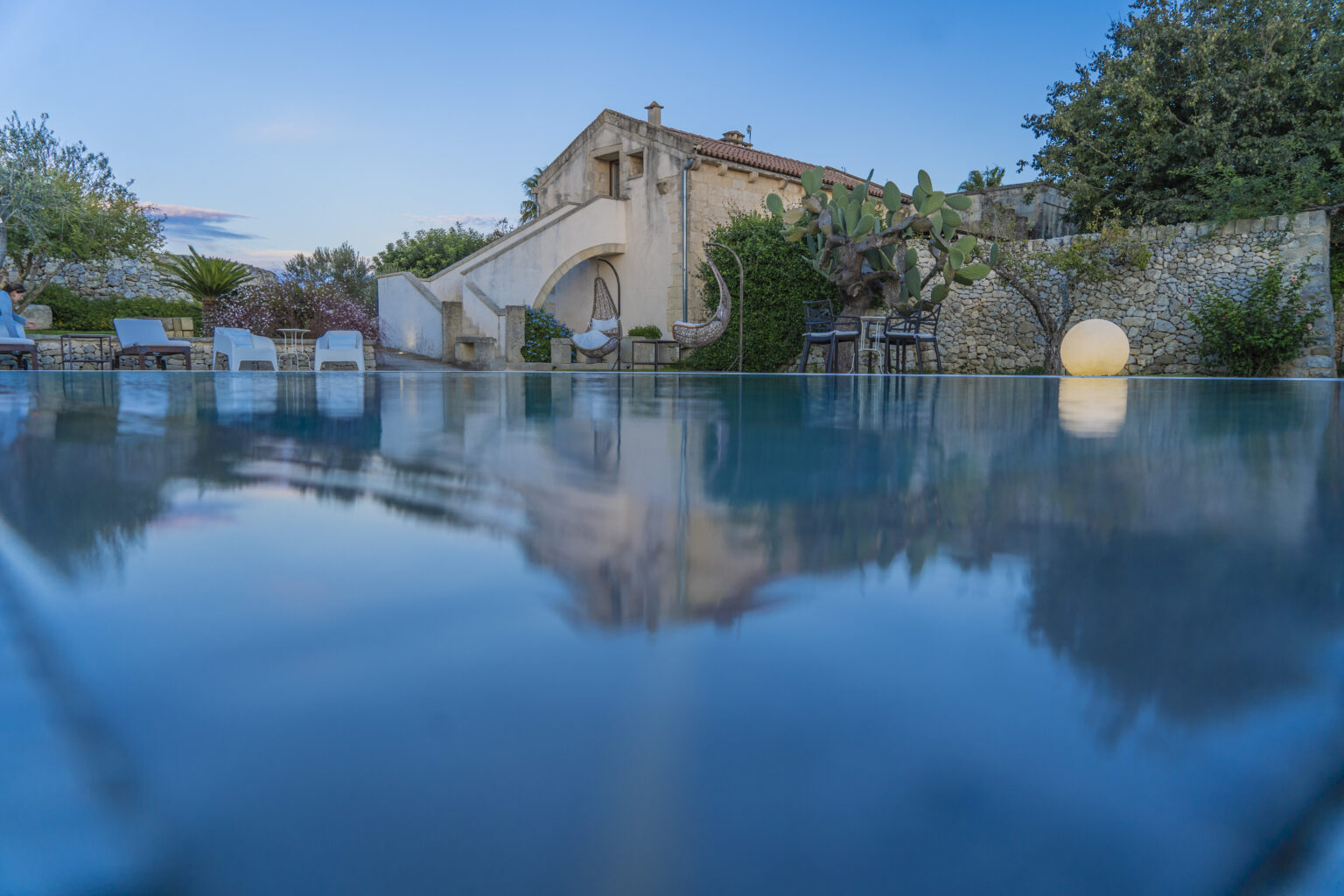 Serene outdoor pool reflecting a charming villa surrounded by lush greenery and modern seating, capturing a tranquil sunset ambiance. | Only on SopranoVillas