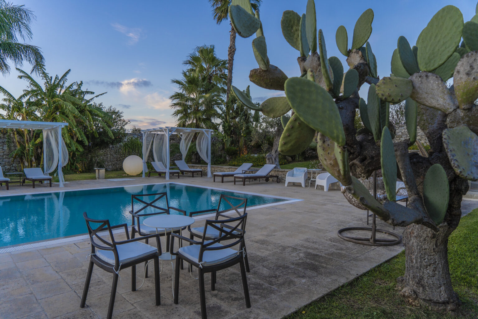 Luxurious outdoor swimming pool surrounded by palm trees and cacti, featuring comfortable lounge chairs and elegant canopies for relaxation, ideal for a serene getaway. | Only on SopranoVillas