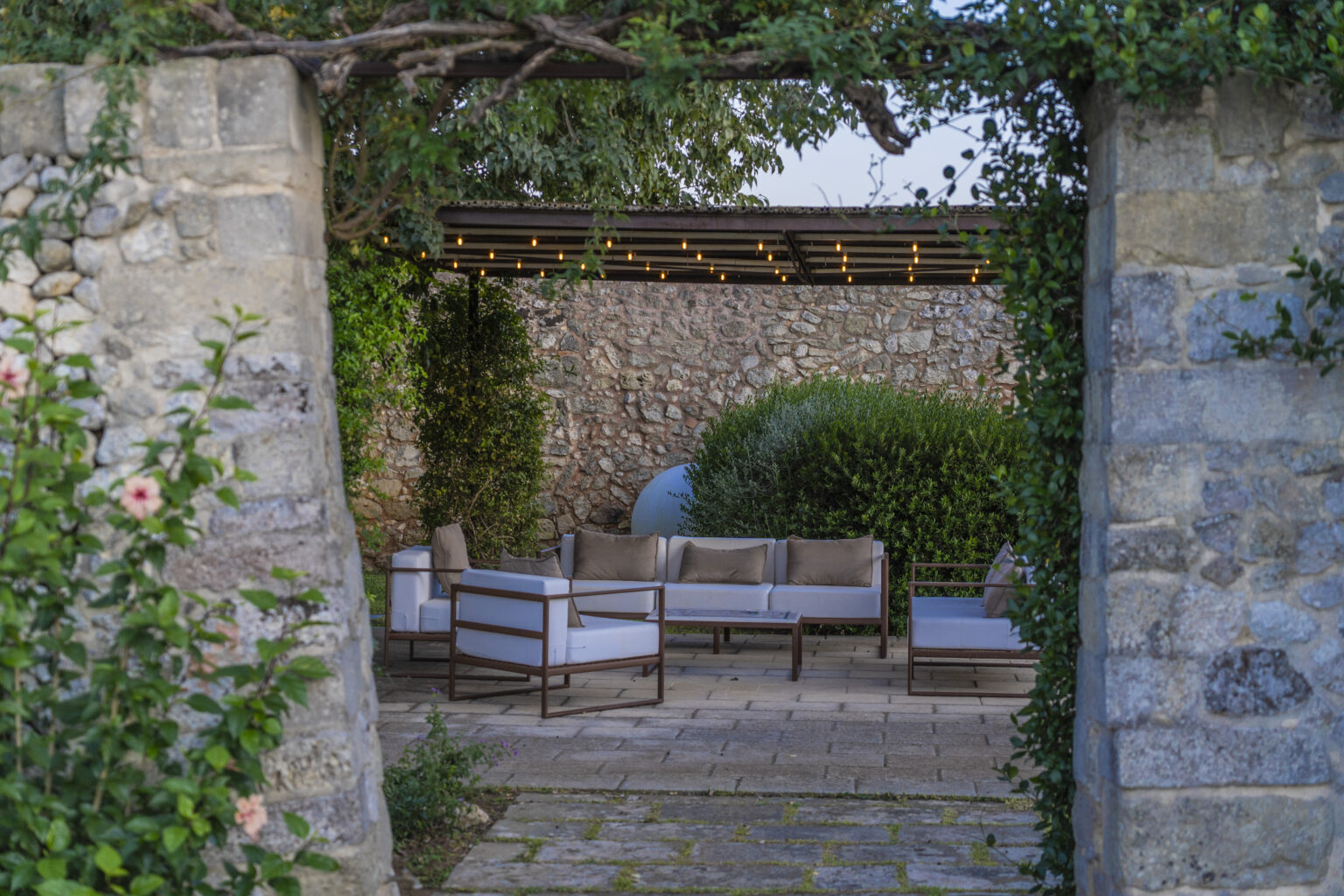 Outdoor seating area framed by stone walls, featuring modern furniture with cushions, surrounded by lush greenery and decorative lighting. Ideal for relaxation and entertaining in a serene garden setting. | Only on SopranoVillas