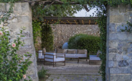 Outdoor seating area framed by stone walls, featuring modern furniture with cushions, surrounded by lush greenery and decorative lighting. Ideal for relaxation and entertaining in a serene garden setting.