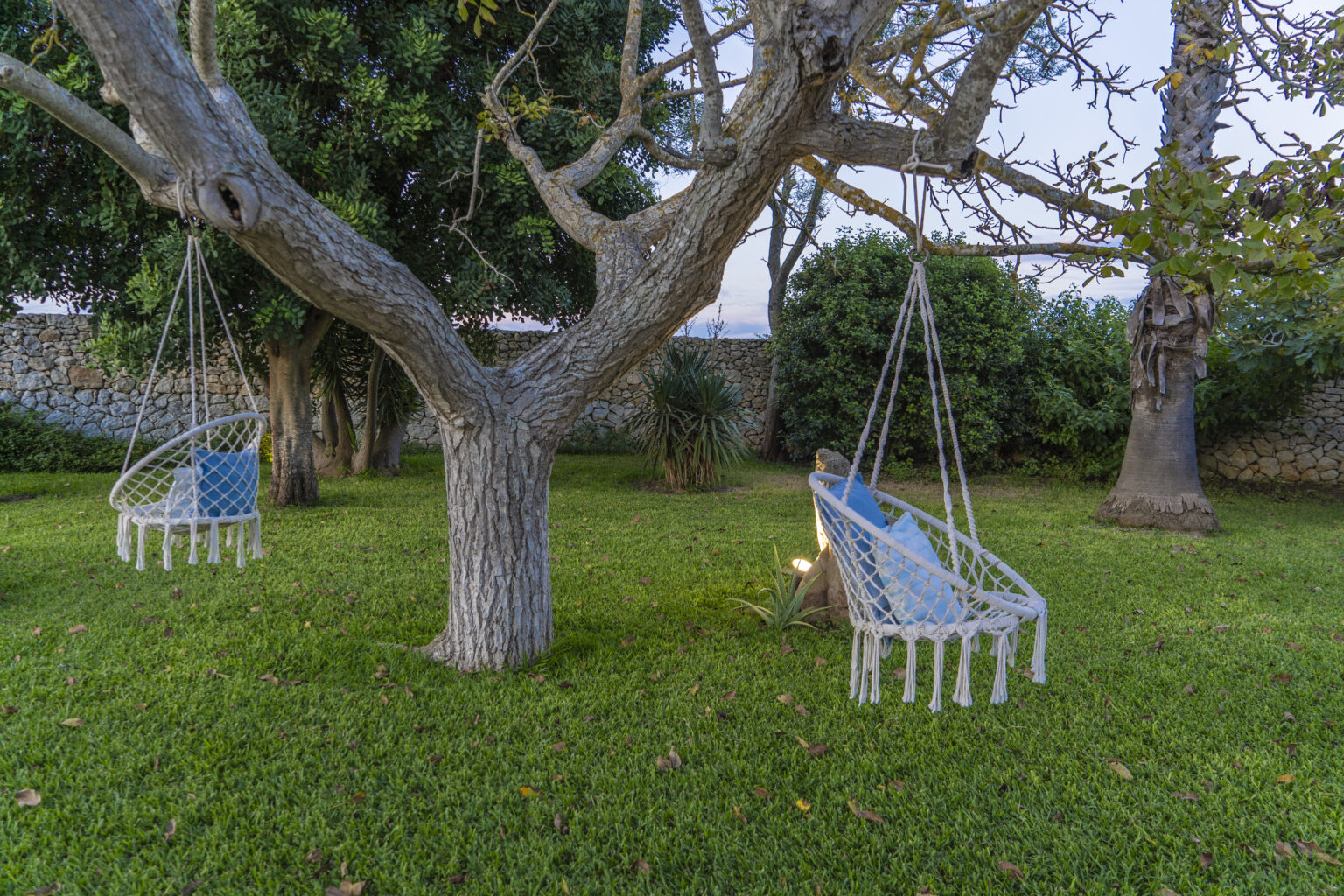 Hammock swings hanging from a tree in a lush green garden, surrounded by various plants and a stone wall in the background, creating a serene outdoor relaxation space. | Only on SopranoVillas
