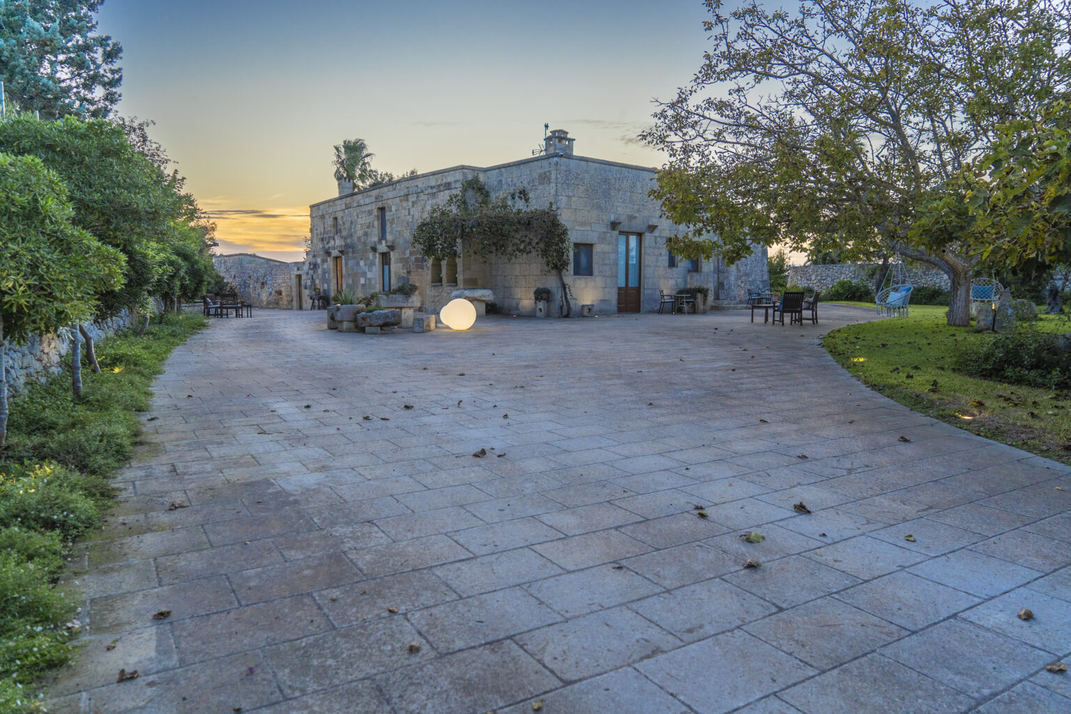 A picturesque outdoor area featuring a stone house, surrounded by greenery and trees, with a spacious paved courtyard illuminated by soft lighting, ideal for relaxation and gatherings at sunset. | Only on SopranoVillas