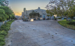 A picturesque outdoor area featuring a stone house, surrounded by greenery and trees, with a spacious paved courtyard illuminated by soft lighting, ideal for relaxation and gatherings at sunset.