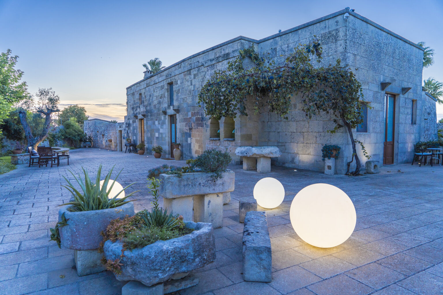 Stone building with lush greenery and decorative glowing orbs in a tranquil outdoor setting at dusk. | Only on SopranoVillas