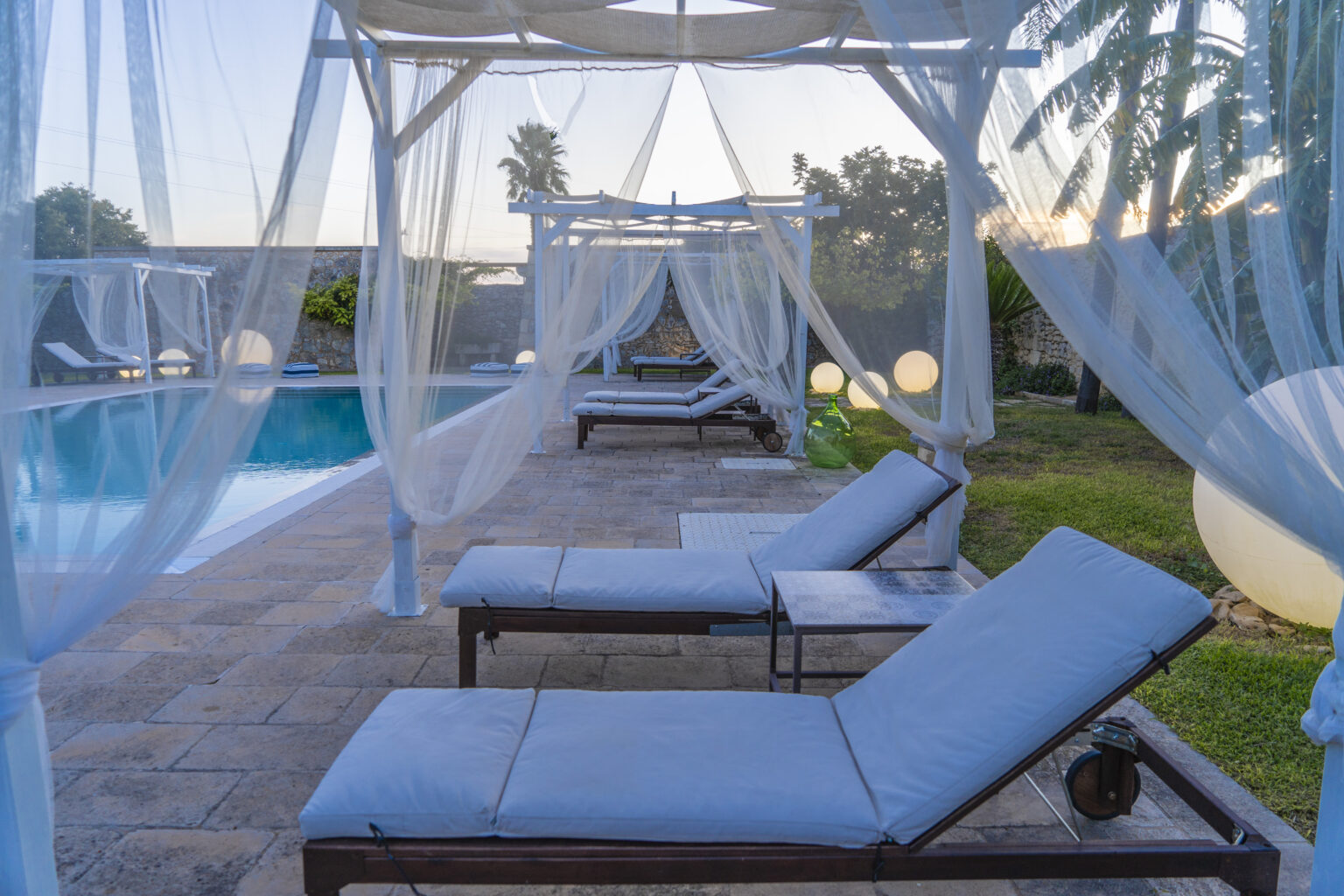 Relaxing outdoor pool area with lounge chairs and sheer canopies, surrounded by lush greenery and palm trees, perfect for leisure and comfort. | Only on SopranoVillas