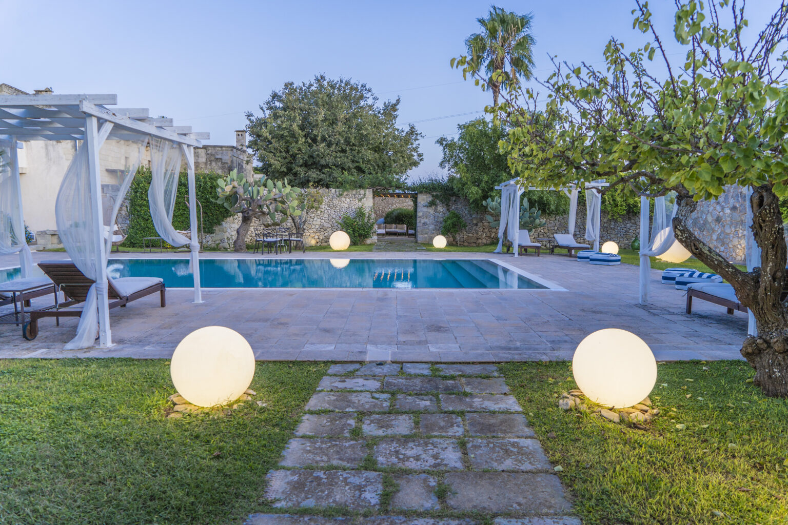 Modern backyard oasis featuring a swimming pool surrounded by lounge chairs, illuminated by decorative globes, with lush greenery and a tranquil atmosphere. Ideal for relaxation and outdoor gatherings. | Only on SopranoVillas