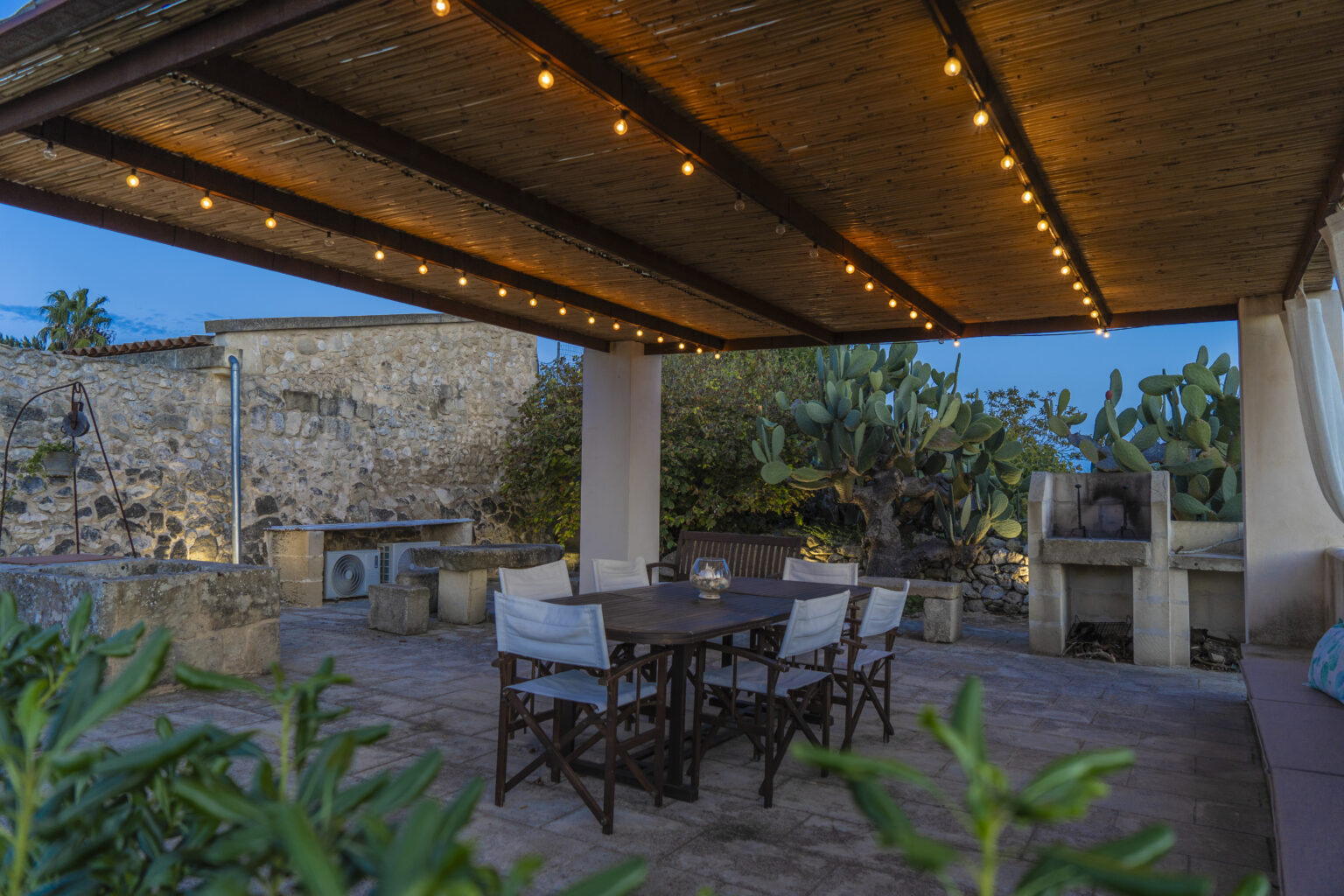 Outdoor dining area with a wooden table and chairs under a pergola, illuminated by string lights, surrounded by cacti and stone walls at dusk. | Only on SopranoVillas