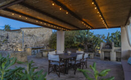 Outdoor dining area with a wooden table and chairs under a pergola, illuminated by string lights, surrounded by cacti and stone walls at dusk.