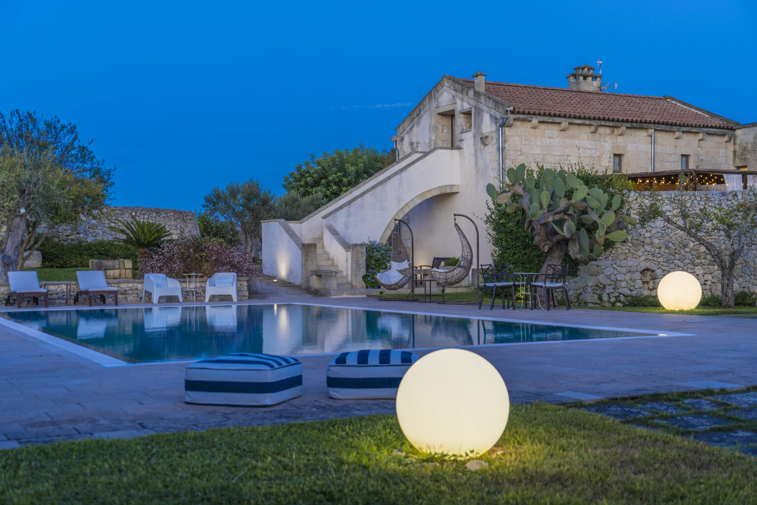 Serene outdoor pool area with modern lighting, featuring a large illuminated sphere, lush greenery, and a rustic stone building in the background, perfect for relaxation and gatherings. | Only on SopranoVillas