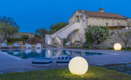 Serene outdoor pool area with modern lighting, featuring a large illuminated sphere, lush greenery, and a rustic stone building in the background, perfect for relaxation and gatherings.