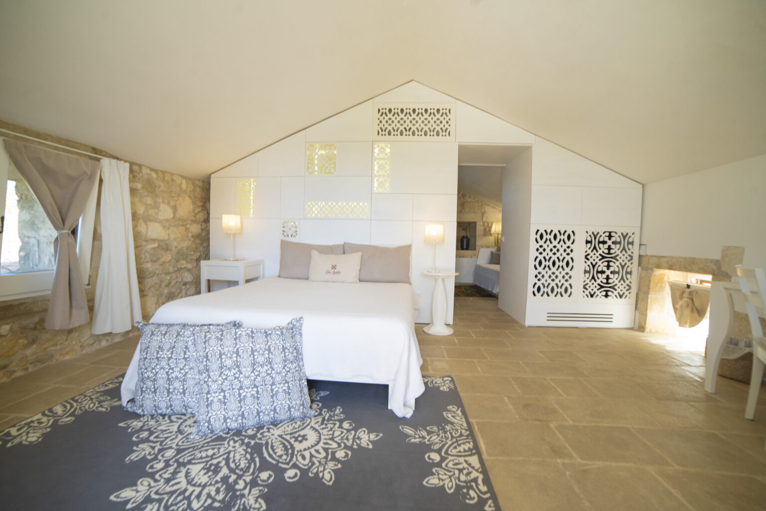 Bright and airy bedroom with a modern design, featuring a white bed, decorative rug, and stylish lighting. Natural stone walls and large windows enhance the tranquil atmosphere, making it an inviting space for relaxation. | Only on SopranoVillas