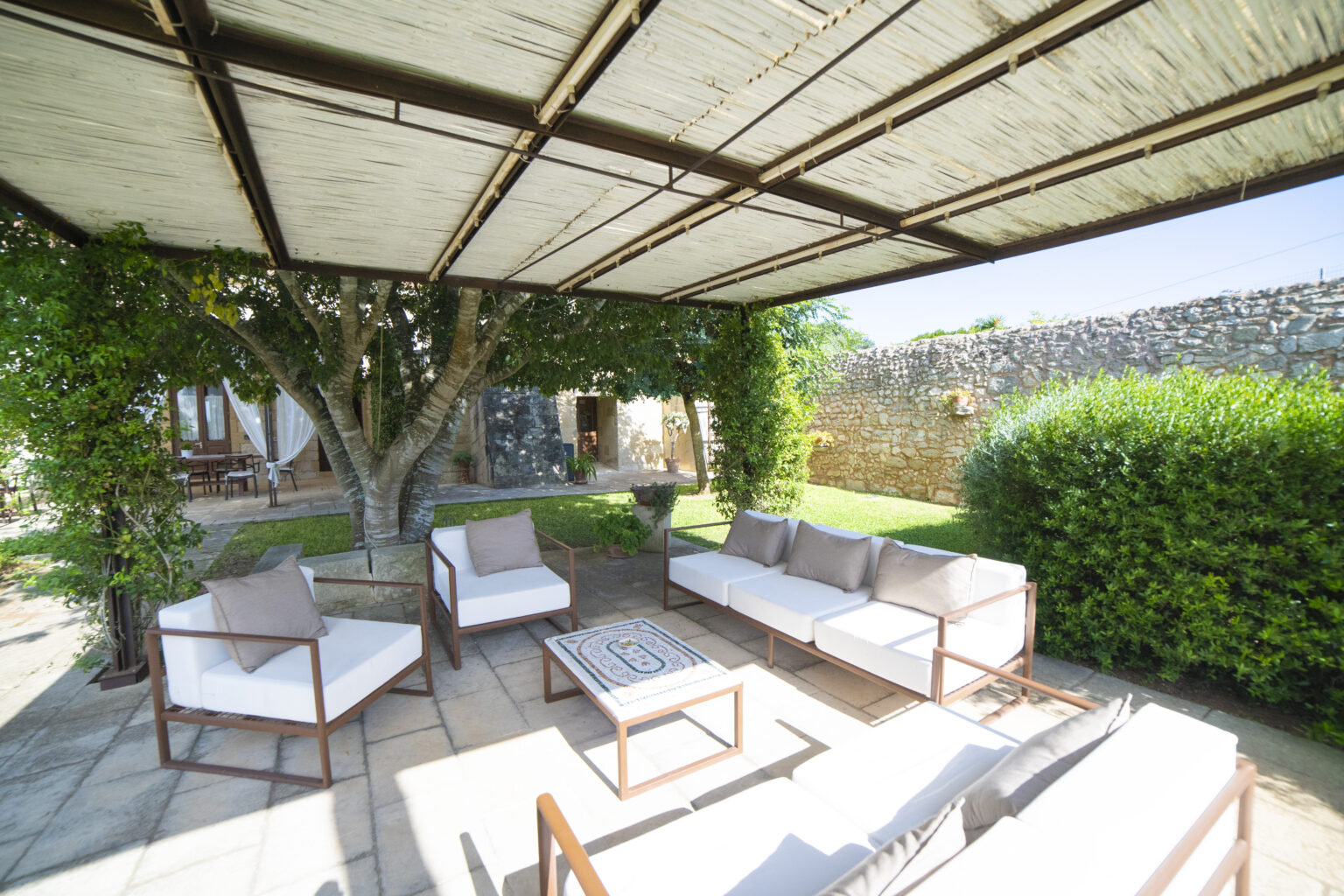 Outdoor patio area featuring comfortable white sofas and a coffee table under a shaded pergola, surrounded by lush greenery and a stone wall, ideal for relaxation and social gatherings. | Only on SopranoVillas
