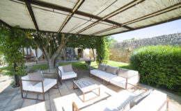 Outdoor patio area featuring comfortable white sofas and a coffee table under a shaded pergola, surrounded by lush greenery and a stone wall, ideal for relaxation and social gatherings.
